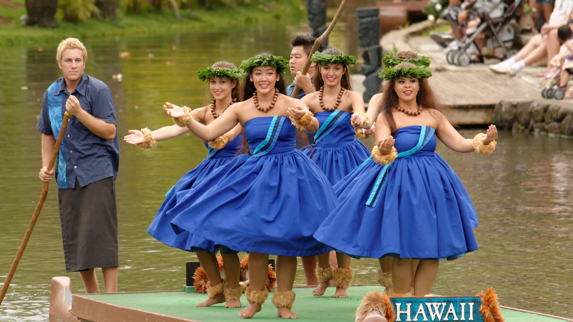 Hula dancers in matching blue dresses performing aboard the “Hawaiʻi” canoe during the Huki canoe pageant on the lagoon at the Polynesian Cultural Center