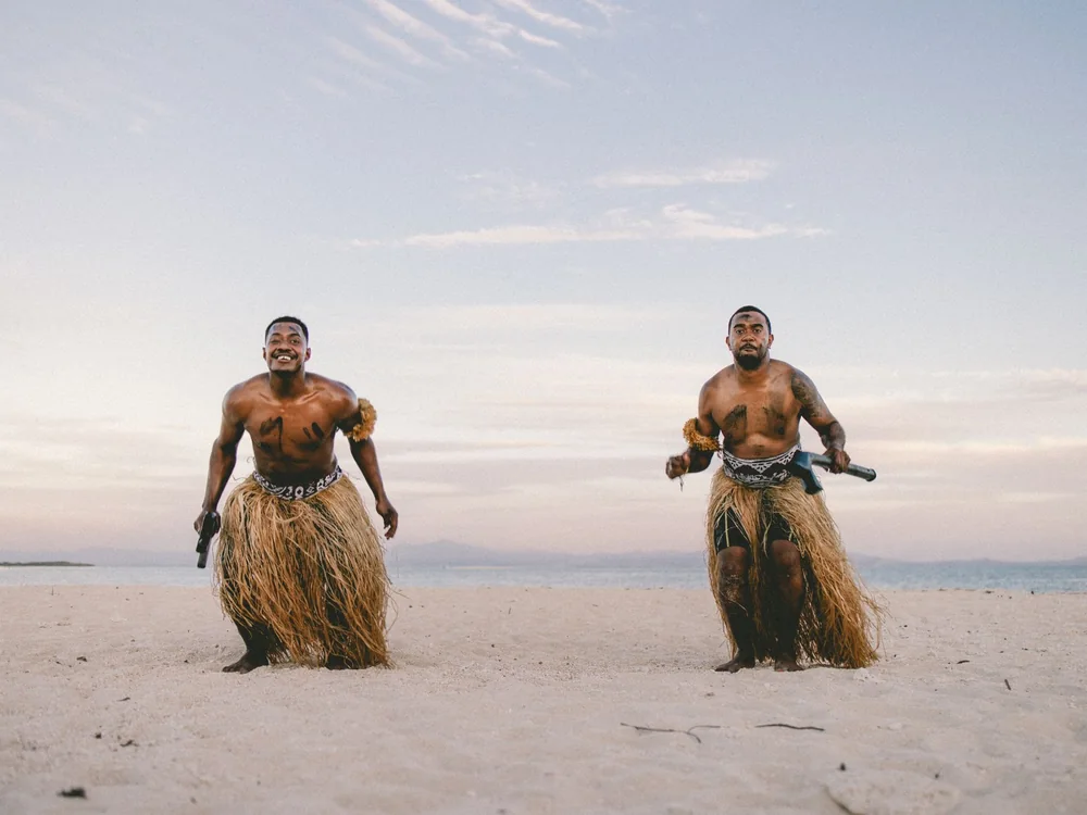 Two men in traditional Fijian dress on a beach at dusk (representative photograph of Fijian dress, not taken at the Center)